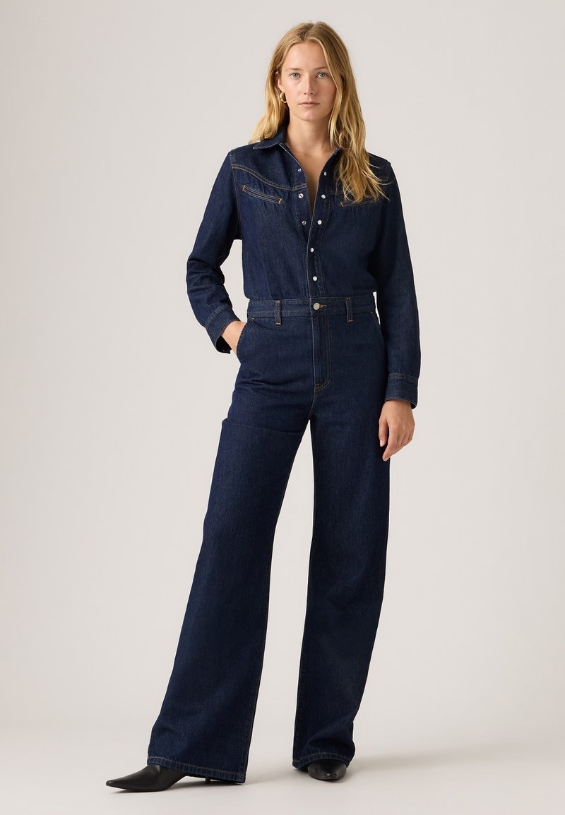 Jeans jumpsuit i mørkeblå farve, med en western-inspireret yoke, knaplukning foran, lange ærmer og bred benudskæring. Tekstureret stof med synlige syninger.
