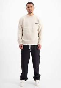 Beige, textuurtrui met geribde manchetten en zoom, met een zwart logo. Zwarte cargo broek met meerdere zakken en witte koord. Witte sneakers.