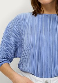 Blaue plissierte Bluse mit einem Schiffsausschnitt, die vertikale Texturlinien und dreiviertel Ärmel aufweist, kombiniert mit einem weißen Taillenband.