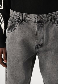 Jeans din denim gri cu un design clasic cu cinci buzunare și închidere cu nasture metalic. Prezentând o textură decolorată și cusături contrastante.