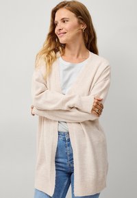 Cardigan beige chiaro realizzato in materiale morbido, caratterizzato da una vestibilità ampia, maniche larghe e un design a V, abbinato a una camicia azzurra chiara e jeans di denim.