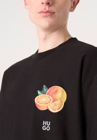 Juodas marškinėliai su spalvinga vaisių grafika, įskaitant visus ir supjaustytus apelsinus, ir balta prekės ženklo tekstu "HUGO" apačioje.
