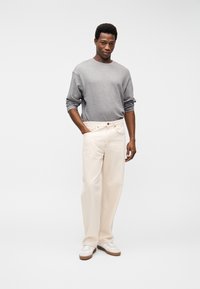 Άνδρας που στέκεται με το ένα χέρι στην τσέπη, φοράει γκρι μακρυμάνικη μπλούζα, κρεμ παντελόνι με φαρδύ πόδι και λευκά αθλητικά παπούτσια, σε λευκό φόντο.