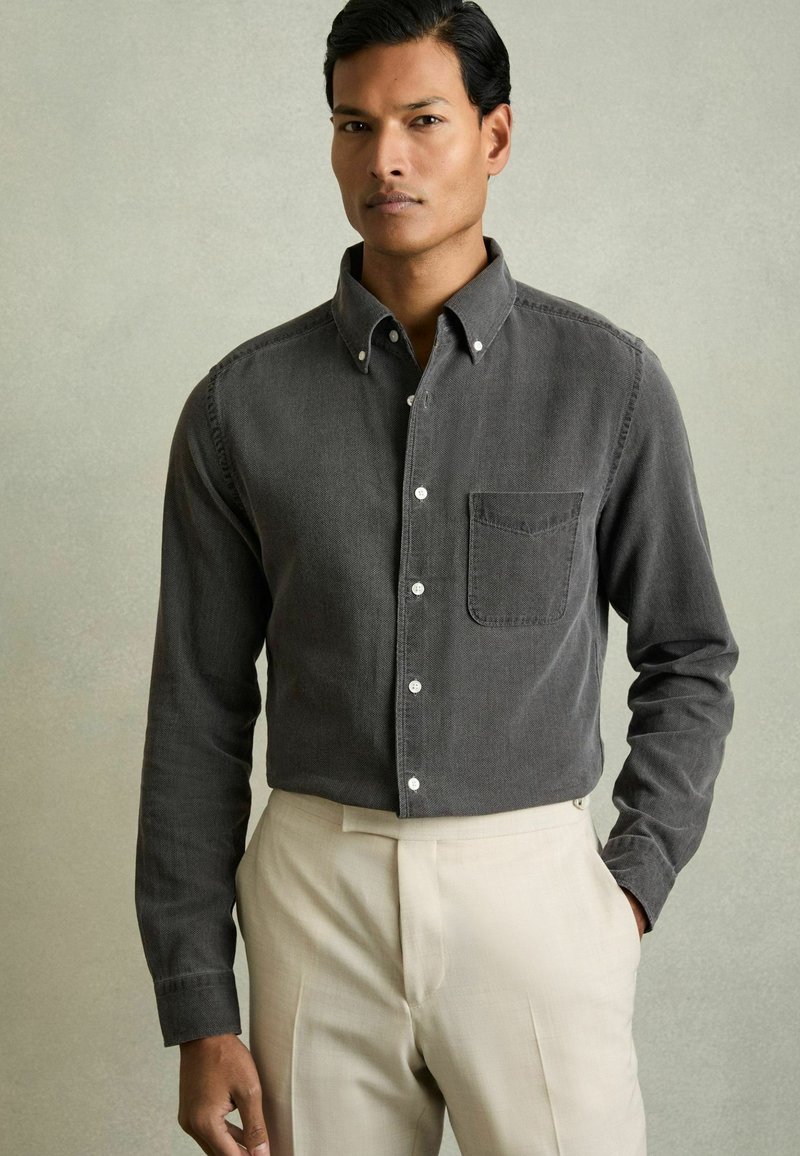 Kolenknop shirt met een borstzak, lange mouwen en witte knopen, gecombineerd met crèmekleurige broek. Subtiele textuur zichtbaar.