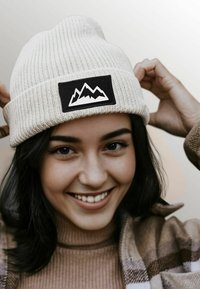 White knit beanie with a folded cuff featuring a black rectangular patch displaying a white mountain design. Soft texture, casual design.