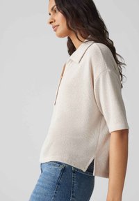 Helle beige Kurzarmbluse mit Kragen, strukturiertem Stoff und seitlichen Schlitzen, kombiniert mit blauen Jeans.