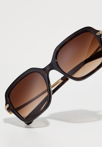 Occhiali da sole oversize con montatura in acetato tartarugato scuro, lenti marroni e dettagli metallici dorati sulle tempie.