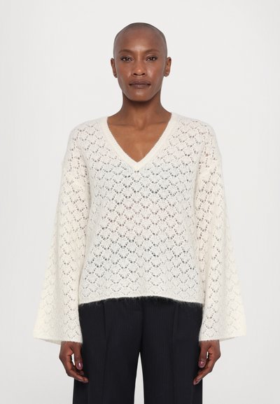 Pull blanc en tricot avec un col en V et des manches évasées. Présente une texture en dentelle et une apparence légèrement transparente. Associé à un pantalon noir à jambes larges.