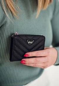 Black quilted wallet made of textured material, featuring a silver zipper and logo accent. Hand with red nails holding the wallet against a green top.
