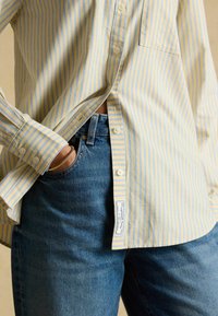 Persona con una camisa de botones de rayas amarillas y azules, ligeramente metida dentro de unos vaqueros azules, con una mano en el bolsillo.