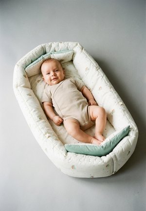 Lächelndes Baby liegt in einer gepolsterten, ovalen Wiege mit leicht gemustertem Stoff und einem kleinen grünen Kissen auf einer grauen Oberfläche.