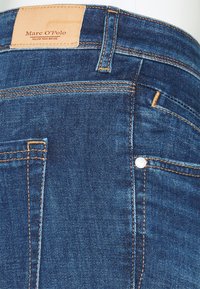 Jean de mezclilla azul con un parche de cuero marrón claro, costuras naranjas y bolsillos frontales. Presenta un pequeño detalle de remache metálico.