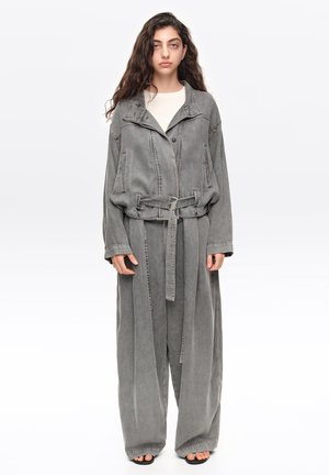 Joven mujer con cabello largo y oscuro lleva una chaqueta gris descolorida y extragrande y pantalones anchos con cinturón, de pie frente a un fondo blanco liso.