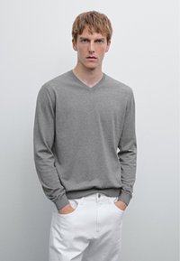 Grauer langärmliger V-Ausschnitt-Pullover aus einem weichen Material, kombiniert mit weißen Hosen mit Seitentaschen und einem knopfverschluss.