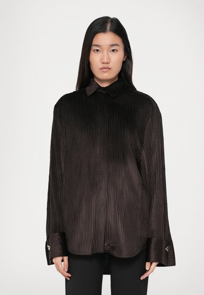 Young woman with long black hair wearing oversized black ribbed shirt and black pants, facing forward with neutral expression.