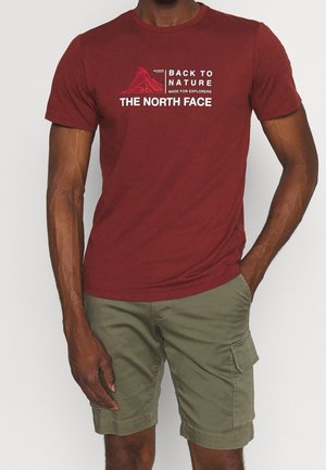 Mężczyzna ubrany w bordową koszulkę North Face z grafiką góry i khaki cargo szorty, stoi z jedną ręką w kieszeni.