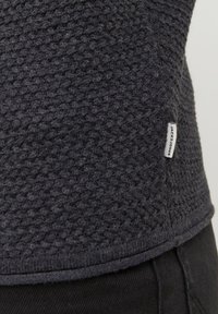 Ciemnoszara, teksturowana dzianinowa bluza z małą białą metką marki "Jack & Jones" przyszytą blisko dolnej krawędzi, noszona na czarnych spodniach.