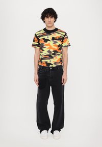 Korte mouwen t-shirt met camouflagepatroon in oranje, groen en zwart, met het woord "BLUE" in zwarte letters, gecombineerd met losse zwarte spijkerbroek en witte sneakers.