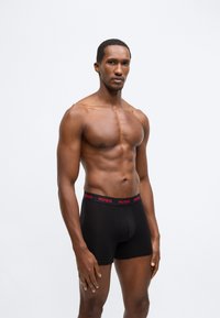 Melnas bokseršorti ar sarkanu "HUGO" jostas daļu, izgatavoti no elastīga auduma, ar pieguļošu dizainu un gludu tekstūru.