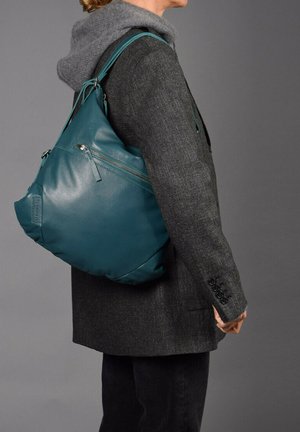 Sac en cuir teal avec une texture douce, des poches zippées et des bretelles réglables. Porté avec un blazer gris et un hoodie.
