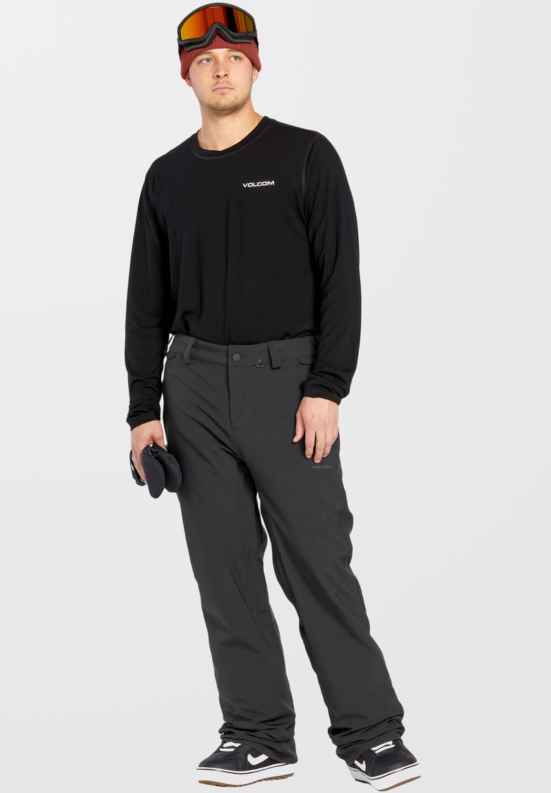 Chemise noire à manches longues avec un logo blanc, associée à un pantalon gris foncé ample. Un homme tient des gants de snowboard; il porte des lunettes de protection et un bonnet.