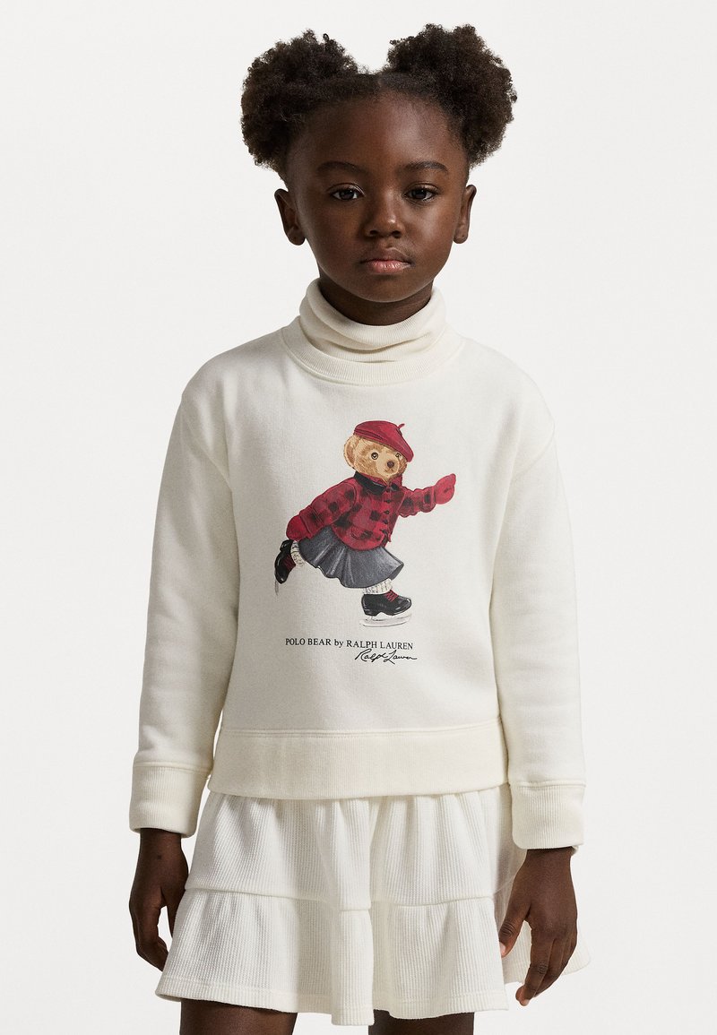 Balta džemperis ar augstu apkakli, ar grafiku, kurā attēlots lācis sarkanas šķērsgriezuma krekliņā un svārkos. Valkāts pāri slāņotām baltām svārkiem.