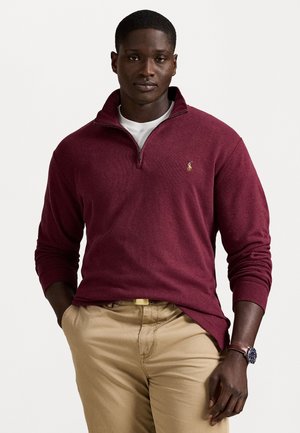 Man met een bordeaux kwart-zip pullover over een wit shirt, beige broek en een bruine horloge, staande met een hand in zijn zak.