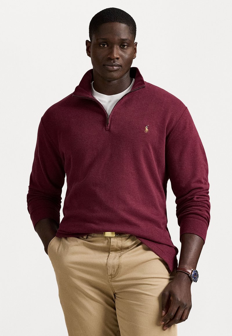 Man wearing a burgundy quarter-zip pullover over a white shirt, beige pants, and a brown watch, standing with one hand in pocket.