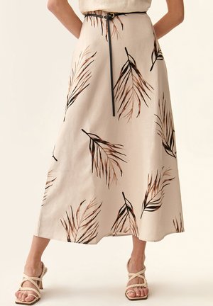 Jupe beige longueur cheville avec des motifs de feuilles noirs et marron, dotée d'une ceinture noire et d'un design fluide. Portée avec des talons beiges à lanières.