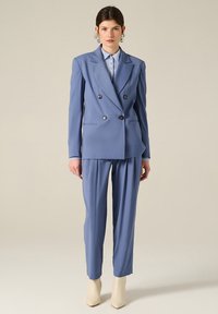 Blazer doppiopetto in tessuto azzurro chiaro con due bottoni scuri. Pantaloni plissettati abbinati e stivaletti alla caviglia color crema.
