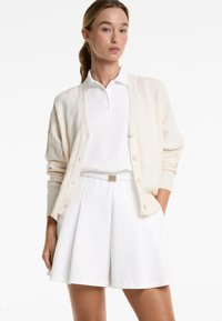 Cream knit cardigan with a diamond pattern, featuring button closures. Worn over a white polo shirt and paired with a white flared skirt.