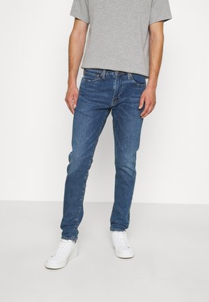 Jeans de mezclilla azul con corte slim, leve desgaste, cinco bolsillos y una cinturilla de altura media, combinados con una camiseta gris y zapatillas blancas.