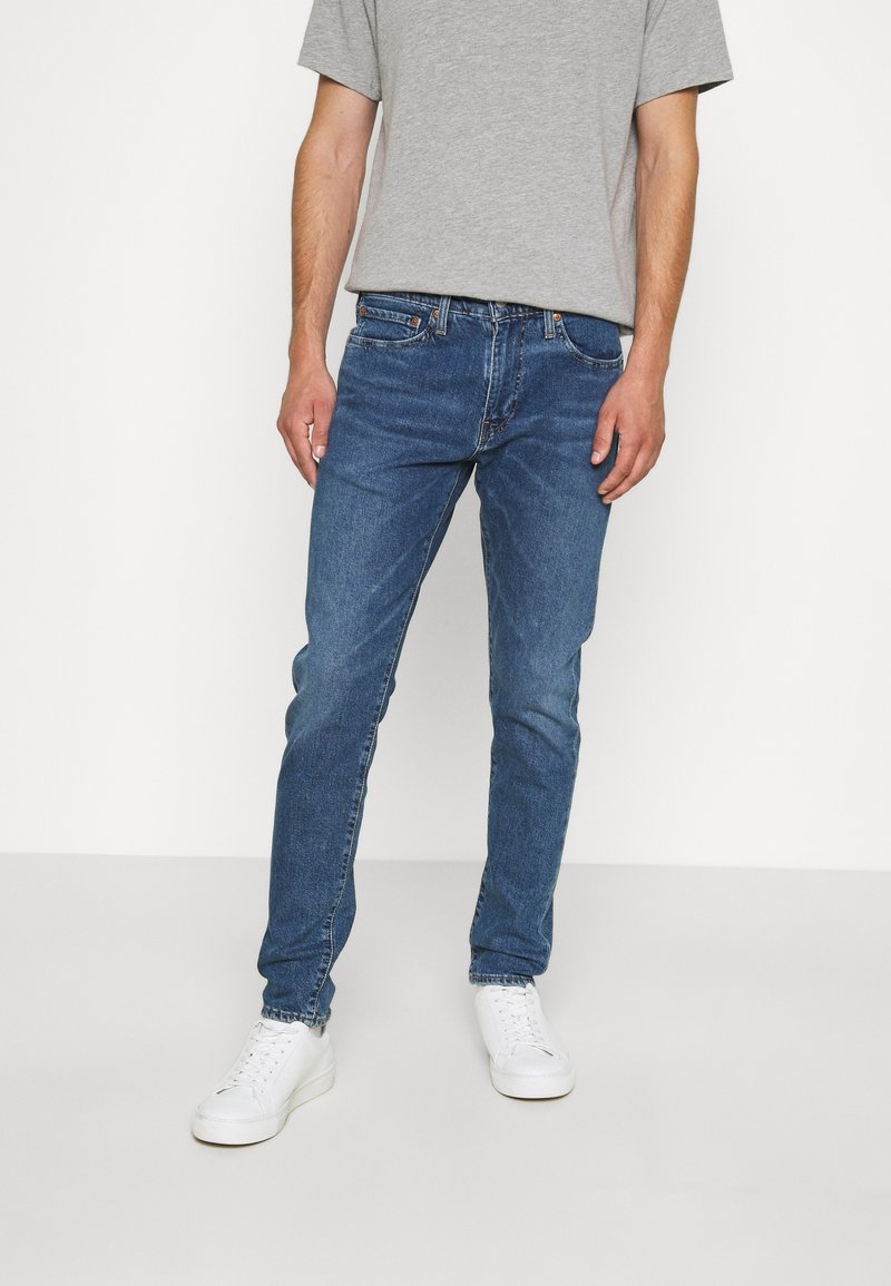 Blå denimjeans med slim passform, lätt blekning, fem fickor och midjehögt linning, matchade med en grå t-shirt och vita sneakers.
