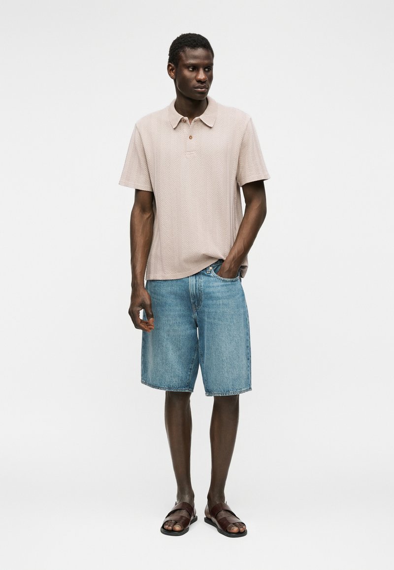 Homme debout, main dans la poche, portant un polo beige à manches courtes, un short en denim bleu clair et des sandales marron sur fond blanc.