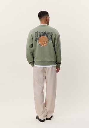 Zielona bluza z grafiką koszykówki i napisem na plecach, w połączeniu z luźnymi beżowymi spodniami i ciemnym obuwiem.