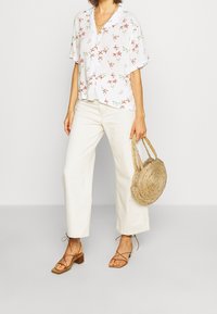 Femme portant un haut blanc à fleurs à manches courtes, un pantalon large blanc cassé, des sandales à talons strappy marron, tenant un sac à main tressé rond.