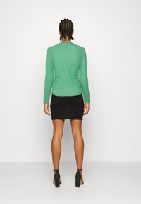 Blusa verde de manga longa combinada com uma saia mini preta. A blusa apresenta uma textura canelada, enquanto a saia é lisa. Sapatos de salto alto pretos.