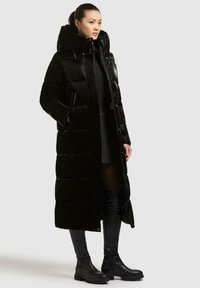 Giacca a vento nera con colletto alto e texture lucida. Presenta un design lungo e aderente con tasche frontali e chiusura con zip.