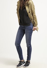 Veste beige avec épaules texturées, haut noir, jean skinny bleu et chaussures de sport noires à lacets. Le mannequin est debout avec une main dans la poche.