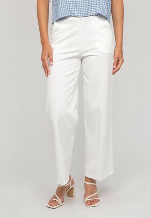 Femme portant un pantalon blanc taille haute à jambes larges, un haut à carreaux bleu, et des sandales blanches à talons blocs avec brides, debout devant un fond uni.