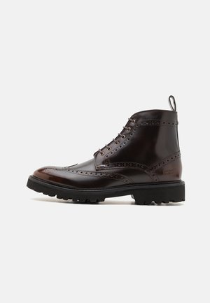 Bottine en cuir marron avec détails brogue, finition lisse, bout rond et semelle épaisse en caoutchouc noir. Comprend une languette à deux boucles.