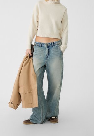 Personne portant un pull à col roulé crème, un jean bleu clair ample, des baskets marron et tenant une veste beige dans une main.