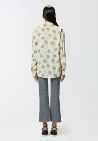 Blusa crema a maniche lunghe con motivi neri ondulati, abbinata a jeans grigi a campana. I dettagli notevoli includono un colletto a ribattina e polsini con bottoni.