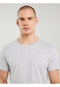 Ung mand med kort buzz-cut hår iført en lys grå t-shirt med rund hals, vendt mod kameraet mod en ensfarvet, lys baggrund.