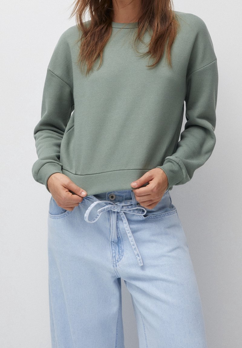 Personne portant un sweat-shirt vert sauge et un jean taille haute bleu clair noué d'un nœud à la taille, debout devant un fond uni.