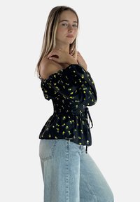 Sort off-the-shoulder bluse med gult blomstermønster, smocket bærestykke og bånd i taljen, parret med lyseblå vide jeans.