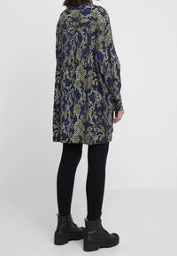 Abito lungo a fantasia in blu scuro, oro e bianco con una vestibilità ampia, maniche lunghe e un tessuto leggermente testurizzato. Leggings neri a costine e stivali neri chunky.