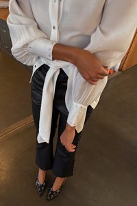 White blouse with buttoned cuffs, tied at the waist. Black faux leather pants with studded accents. Black pointed shoes with silver details.