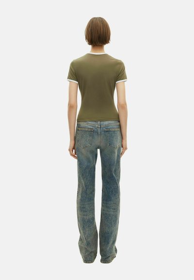 Person vist fra bagfra iført en olivengrøn kortærmet skjorte med hvid kant og falmede blå jeans mod en ensfarvet hvid baggrund.