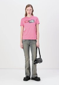 Rosa t-skjorte med korte ermer og en svart-hvitt Diesel-logo, sammen med lysegrå vidde jeans og svarte platå sko, holder en liten svart veske.